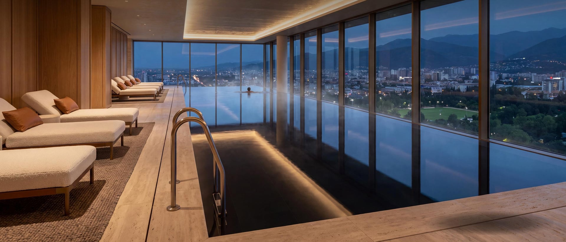 An indoor pool at dusk running along full-height glazing, loungers with rolled towels on a timber deck, and the lit city and the mountains beyond.