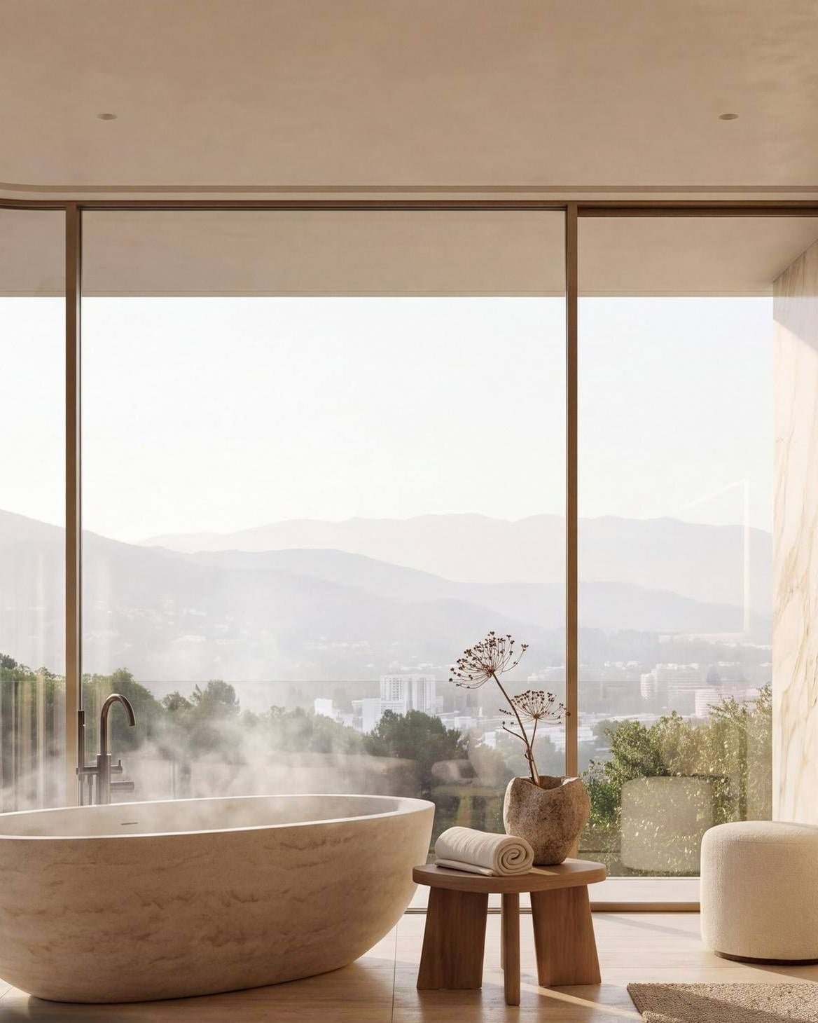 A principal bathroom in travertine, a freestanding stone bath steaming at the full-height glass, with the valley and the mountains beyond.